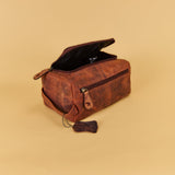 A brown distressed leather travel pouch with an open top zipper and a hanging leather tassel, shown on a solid beige background.