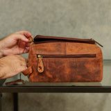 A person's hands holding open a brown distressed leather zippered pouch or toiletry bag, resting on a dark glass table against a textured gray concrete wall.