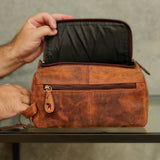 A person's hands open a brown distressed leather toiletry bag, showing its black interior lining and zipper compartment, set on a glass table against a textured gray wall.