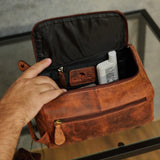 A brown distressed leather travel toiletry bag shown open on a glass table, with a hand holding it and a white bottle inside.