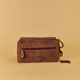 A brown distressed leather travel pouch with a front zippered pocket and a small hanging leather tag, set against a solid beige background.