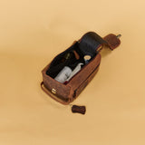 A brown leather travel or wine bag with an open flap, showing a dark interior holding several bottles, set against a solid beige background.