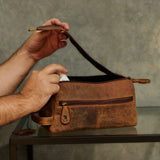 A person's hands are shown holding open a distressed brown leather travel toiletry bag, displaying its interior. The bag is resting on a dark glass table against a textured gray concrete wall.