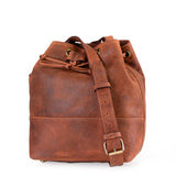 The Rosa Leather Crossbody Bag by Vintage Leather Sydney is a brown leather bucket bag with a long adjustable strap, gold-tone buckle, and drawstring closure. Spacious and stylish, it stands upright against a white background.