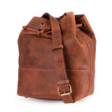 The Rosa Leather Crossbody Bag by Vintage Leather Sydney is a brown leather drawstring bucket bag with a long adjustable shoulder strap and gold-tone buckle, shown against a white background.