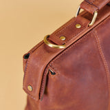 Close-up of a brown leather bag showing a strap with brass-tone rivets and a D-ring closure against a warm beige background.