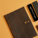 The Vintage Leather Oxford Leather A4 Notebook Cover, in brown with a snap closure, sits with a black pen in a box and another item on a light tan surface—an elegant choice for daily notes.