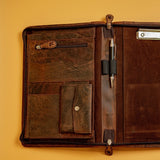 The Vintage Leather Sydney A4 Leather Compendium - Cairo is shown open on a light yellow surface, featuring durable brown leather, multiple pockets, a zippered pouch, snap-button pocket, pen slot with pen, and a right-side clipboard.