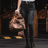A person in black jeans and boots holds the Leather Weekender Bag - Altico by Vintage Leather, standing in front of a fruit market display with colorful apples and oranges in the background.