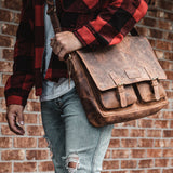 A person in a red plaid jacket and ripped jeans holds the Vintage Leather Easton Leather Messenger Bag, featuring buckles and a padded laptop compartment, while standing against a brick wall.