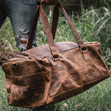 A person in ripped jeans holds the Vintage Leather Weekender Bag in Texas Brown, featuring sturdy straps and a distressed finish, standing on grass with greenery behind.