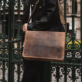 A person in a black jacket and dark pants stands before an ornate green metal gate, carrying the Vintage Leather Jersey Brown Laptop Bag for men over their shoulder.