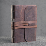 The Indigo Journal Leather by Vintage Leather features brown buffalo leather with visible spine stitching and a long wraparound strap, standing upright on a gray textured surface—ideal for personalization.