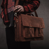 Wearing a red plaid jacket and graphic tee, a person holds the Vintage Leather Messenger Bag - Toledo by Vintage Leather. The plain background accentuates its vintage style, multiple pockets, and sturdy shoulder strap.