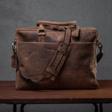 The Vintage Leather Colombo Laptop Bag, crafted from brown full-grain leather with a shoulder strap and vintage design, sits on a wooden surface against a gray backdrop. It features a rugged look and a dedicated laptop compartment.