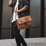 A person in a black denim jacket, white shirt, and black pants stands on steps with the Vintage Leather Melbourne Leather Messenger Bag, featuring buckle details and a spacious laptop compartment. Modern building windows are in the background.