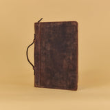 A dark brown distressed leather journal with a black leather strap closure, standing upright on a warm beige background.