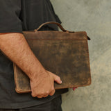 A person in a black shirt holds a distressed brown leather briefcase against their side, set against a textured gray concrete wall.