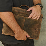 A person in a black shirt holds a distressed brown leather laptop sleeve or briefcase with a top handle and front pocket, shown against a gray concrete wall.