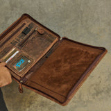 A brown leather organizer case shown open, held by a person whose arm is visible on the left. The case contains multiple card slots, a phone pocket with a blue screen protector, and a zippered compartment.