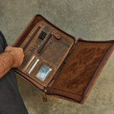 A brown leather organizer case shown open, held by a person whose arm and dark shirt are partially visible against a textured gray concrete background. The case features multiple interior card slots, a black pen loop, and a zippered main compartment.