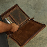 A person holding open a brown leather portfolio or organizer case, showing interior card slots and pockets, against a textured gray concrete background.