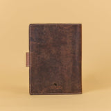 A dark brown distressed leather notebook cover or planner sleeve with a small leather tab on the side and a debossed logo near the bottom, set against a solid beige background.