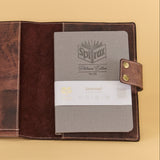 A brown leather journal cover shown open on a warm beige background, holding a gray hardcover journal with a white paper band and a blue pen in a side pocket.