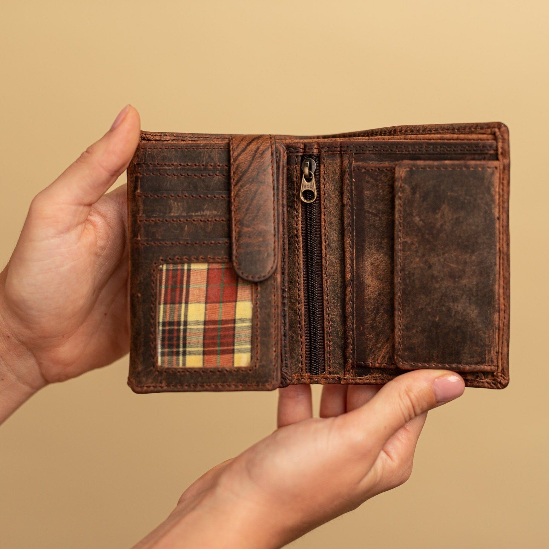 Two hands hold the Vintage Leather Sydney Bento women's leather wallet, featuring card slots, a clear ID window with plaid pattern, zippered pocket, and coin pouch—stylish and compact for everyday use against a beige background.
