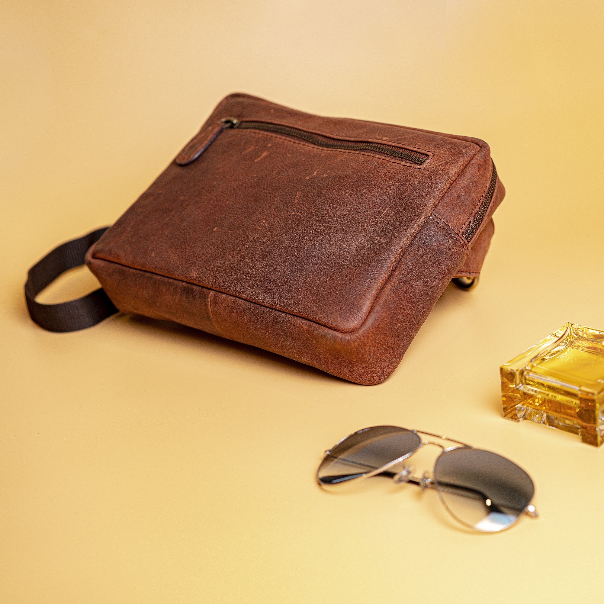 The Leather Sling Bag Akiva by Vintage Leather Sydney, in brown leather with a front zipper pocket, rests on a light yellow surface next to aviator sunglasses and a square glass perfume bottle.