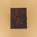 A dark brown leather journal with a matching wrap-around strap and a small metal snap button, displayed flat against a solid beige background.