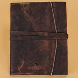 A dark brown distressed leather notebook or journal with a matching elastic band closure and white stitching, featuring an embossed logo at the bottom, set against a solid beige background.