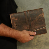 A person in a black shirt holds a distressed brown leather folio or notebook cover with a strap closure and white stitching against a textured gray background.