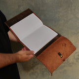 A person holding an open brown leather notebook or journal with lined white pages, shown against a textured gray concrete wall.