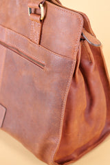 A close-up of a brown leather handbag showing its texture, stitching, zipper pocket, and brass hardware against a warm beige background.