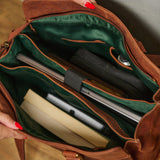 Open brown leather bag showing a green interior organizer with compartments holding a laptop, notebook, and other items. A hand with red nail polish holds the bag open.