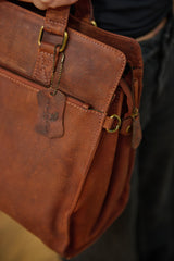 A close-up of a person holding a brown textured leather briefcase or satchel with brass-tone hardware and a hanging leather brand tag.