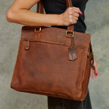 A person holding a large brown leather tote bag with gold-tone hardware and a hanging leather tag, shown against a gray textured background.