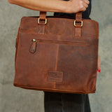 A brown distressed leather tote bag with gold-tone hardware, held by a person against a gray concrete wall.