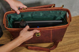 A brown leather briefcase or laptop bag shown open, with a dark green interior lining and a laptop tucked inside. Hands are visible holding the bag open, and the product is photographed on a wooden surface against a neutral gray background.