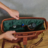 A brown leather bag organizer shown open with hands holding it, revealing a dark green interior organizer pocket holding small bottles and a pen. The bag is photographed indoors on a wooden surface against a gray wall.