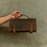 A hand holding a dark brown distressed leather travel tool pouch with brass buckles and a top handle, set against a textured gray background.
