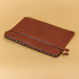A brown leather zippered pouch or sleeve with a small rectangular logo patch on the front, photographed on a warm beige studio background.
