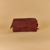 A brown leather zippered pouch with a rectangular embossed logo patch on the front, displayed against a solid beige background.