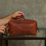 A person's hands are shown holding a brown leather travel or toiletry pouch on a glass-topped table against a textured gray wall.