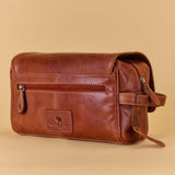 A brown leather travel pouch with a front zippered pocket and a small embossed logo patch, shown on a solid beige background.
