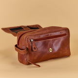 A brown leather toiletry bag with a zippered front pocket and a gold snap button, shown with its flap open on a solid beige background.
