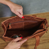 A brown leather tote bag shown open, with two hands holding the top and bottom edges to display the interior compartments, zipper pockets, and lining. The bag is photographed over a wooden surface against a gray wall.