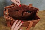 A brown leather tote bag shown open, with two hands holding the zipper area to display the interior organization pockets and contents.