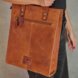 A person carrying a large cognac brown leather tote bag with brass hardware and a front zipper pocket, shown against a neutral gray background.